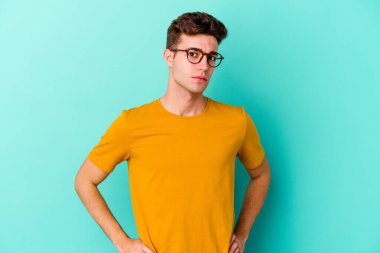 Kafkasyalı genç adam mavi arka planda izole edilmiş hoşnutsuzca kaşlarını çatmış, kollarını kavuşturmuş..