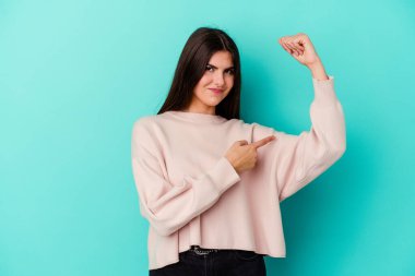 Mavi arka planda izole edilmiş genç beyaz kadın güçlü bir hareket sergiliyor kollarıyla, kadınsı gücün sembolü.