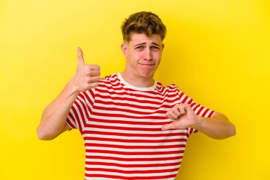 Sarı arka planda izole edilmiş genç beyaz adam baş parmağını kaldırıyor ve baş parmak aşağı, zor seçim konsepti.