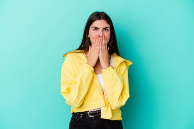 Mavi arka planda izole edilmiş genç beyaz kadın şok olmuş, ağzı ellerle kaplanmış, yeni bir şey keşfetmeye hevesli..