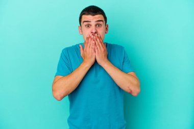 Mavi arka planda izole edilmiş genç beyaz adam elleriyle ağzını kapatırken şok oldu..