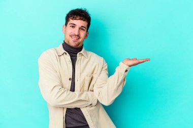 Mavi arka planda izole edilmiş genç beyaz bir adam bir palmiye üzerinde fotokopi alanı ve belinde bir el daha gösteriyor..