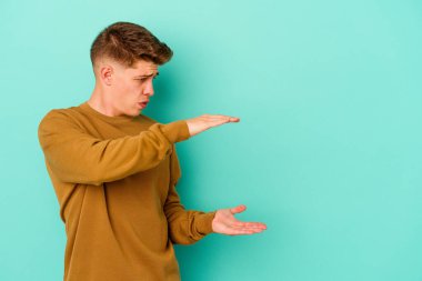 Mavi arka planda izole genç beyaz adam şok ve hayret içinde elleri arasında bir kopya boşluk tutuyor.