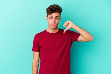 Mavi arka planda izole genç beyaz adam baş parmağını aşağı gösteriyor, hayal kırıklığı konsepti.