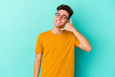 Mavi arka planda izole edilmiş genç beyaz adam parmaklarıyla bir cep telefonu hareketi gösteriyor..