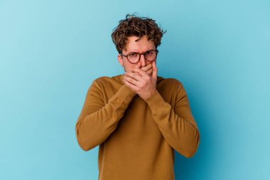 Gözlüklü genç beyaz adam mavi arka planda izole edilmiş ağzı kapalı ve elleri endişeli görünüyor..