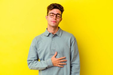 Sarı arka planda izole edilmiş genç beyaz adam karnına dokunur, nazikçe gülümser, yemek yer ve tatmin olur..