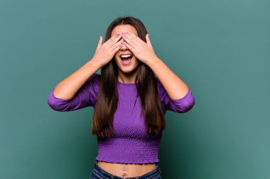 Mavi arka planda soyutlanmış genç bir Hintli kadın elleriyle gözlerini kapatıyor, geniş bir gülümsemeyle bir sürpriz bekliyor..