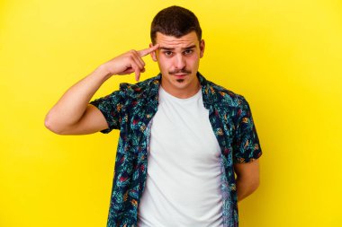 Sarı arka planda izole edilmiş genç beyaz adam şakağı parmağıyla işaret ediyor, düşünüyor ve bir göreve odaklanıyor..