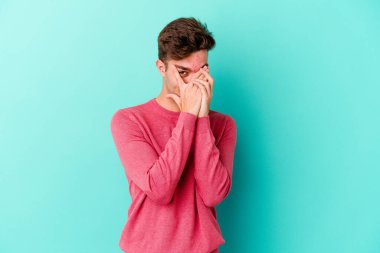 Kafkasyalı genç adam mavi arka planda izole edilmiş. Korkmuş ve gergin parmaklarıyla göz kırp..