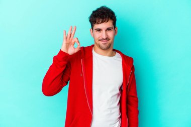 Kafkasyalı genç bir adam mavi arka planda izole edilmiş bir şekilde göz açıp kapayana kadar iyi bir el hareketi yapıyor..