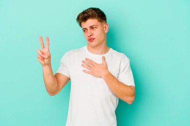 Kafkasyalı genç adam mavi arka planda izole edilmiş yemin ediyor, elini göğsüne koyuyor..