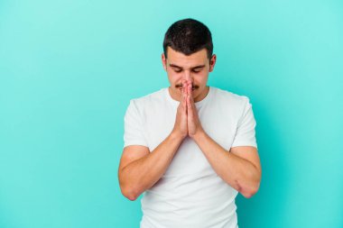Mavi arka planda izole edilmiş genç beyaz bir adam el ele tutuşup, ağzının kenarında dua ediyor, kendinden emin..
