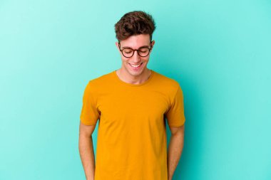 Mavi arka planda izole edilmiş genç beyaz adam gülüyor ve gözlerini kapıyor, rahatlamış ve mutlu hissediyor..