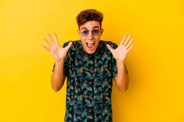 Hawaii gömleği giyen genç beyaz adam sarı arka planda izole bir şekilde zaferi veya başarıyı kutluyor, şaşırıyor ve şok oluyor..