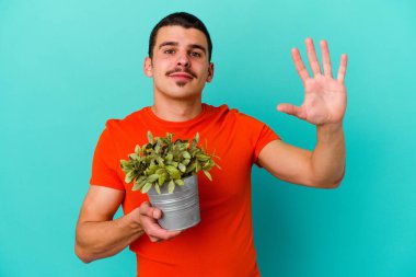 Mavi arka planda yaprak tutan beyaz bir adam gülümseyerek beş numarayı parmaklarıyla gösteriyor..