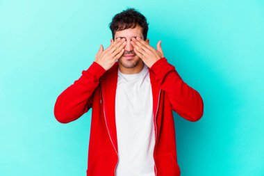Mavi arka planda izole edilmiş genç beyaz adam elleriyle gözlerini kapatmaktan korkuyor..