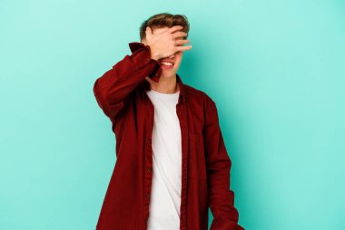 Mavi arka planda soyutlanmış genç beyaz bir adam elleriyle gözlerini kapatıyor, geniş bir gülümsemeyle bir sürpriz bekliyor..