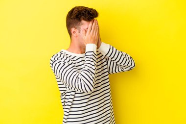 Sarı arka planda izole edilmiş genç beyaz adam elleriyle gözlerini kapatmaktan korkuyor..