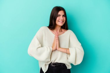 Mavi arka planda izole edilmiş genç beyaz bir kadın el ele tutuşup ağza yakın dua ediyor, kendinden emin..