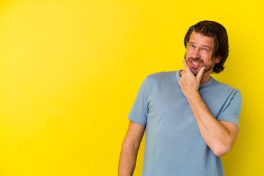 Orta yaşlı beyaz bir adam sarı arka planda izole edilmiş kafasının arkasına dokunuyor, düşünüyor ve bir seçim yapıyor..