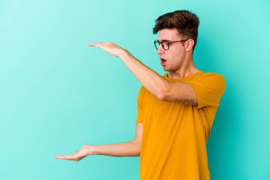 Mavi arka planda izole genç beyaz adam şok ve hayret içinde elleri arasında bir kopya boşluk tutuyor.