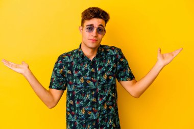 Hawaii gömleği giyen genç beyaz adam sarı arka planda izole edilmiş sorgulama jestinde şüphe ediyor ve omuz silkiyor..