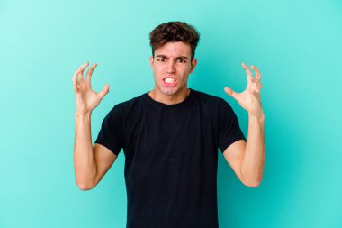 Mavi arka planda izole edilmiş genç beyaz adam öfkeyle bağırıyor..