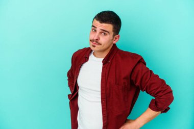 Kafkasyalı genç adam mavi arka planda izole, kafası karışmış, şüpheli ve güvensiz hissediyor..