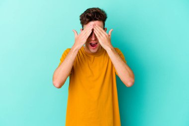 Mavi arka planda soyutlanmış genç beyaz bir adam elleriyle gözlerini kapatıyor, geniş bir gülümsemeyle bir sürpriz bekliyor..