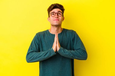 Sarı arka planda izole edilmiş genç beyaz bir adam el ele tutuşup ağız kenarında dua ediyor, kendinden emin hissediyor..