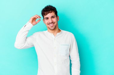 Mavi arka planda izole edilmiş genç beyaz bir adam bir şeye gülüyor, ağızlarını ellerle kapatıyor..