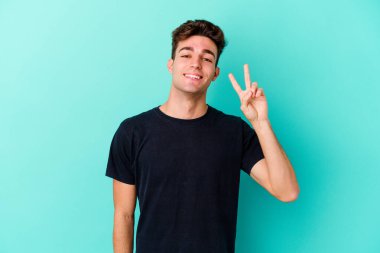 Mavi arka planda izole edilmiş genç beyaz adam zafer işareti gösteriyor ve gülümsüyor..