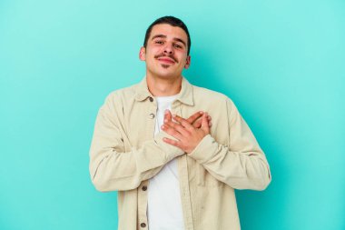 Kafkasyalı genç adam mavi arka planda izole olmuş gülüyor, elini yüreğine koyuyor, mutluluk kavramını koruyor..