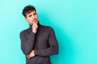 Mavi arka planda izole edilmiş genç beyaz adam şüpheli ve şüpheci bir ifadeyle yana bakıyor..