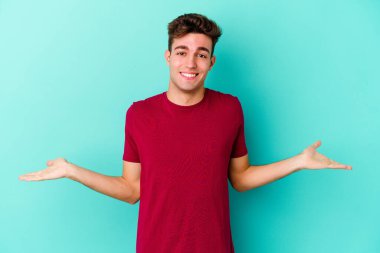 Mavi arka planda izole genç beyaz adam kollarıyla ölçek yapar, mutlu ve kendinden emin hisseder..