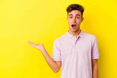 Sarı arka planda izole edilmiş genç beyaz adam, avuç içi fotokopi alanını elinde tutmaktan etkilendi..