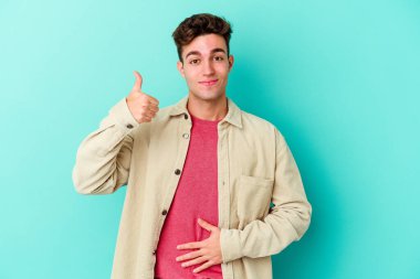 Mavi arka planda izole edilmiş genç beyaz adam karnına dokunuyor, nazikçe gülümsüyor, yemek yiyor ve tatmin oluyor..