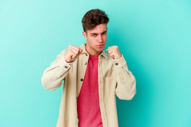 Kafkasyalı genç bir adam mavi arka planda izole edilmiş. Yumruk atıyor, sinirleniyor, kavga ediyor, kavga ediyor..