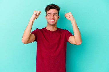 Kafkasyalı genç adam mavi arka planda izole edilmiş kaygısız ve heyecanlı tezahüratlar yapıyor. Zafer konsepti.
