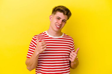 Sarı arka planda izole edilmiş genç beyaz adam iki parmağını da kaldırıyor, gülümsüyor ve kendinden emin..