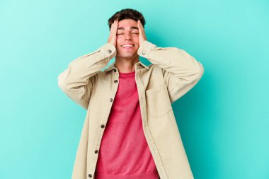 Mavi arka planda izole edilmiş genç beyaz adam ellerini başının üstünde tutarak neşe içinde gülüyor. Mutluluk kavramı.