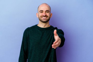 Mor arka planda izole edilmiş, genç, beyaz, kel bir adam. Kameraya doğru elini uzatarak selam veriyor..