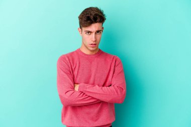 Kafkasyalı genç adam mavi arka planda izole edilmiş hoşnutsuzca kaşlarını çatmış, kollarını kavuşturmuş..