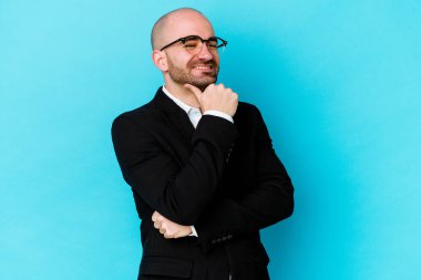 Genç beyaz tenli kel adam mavi arka planda izole edilmiş mutlu ve kendinden emin gülümsüyor, elleriyle çenesine dokunuyor..