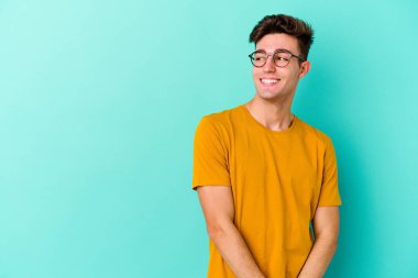 Mavi arka planda izole edilmiş genç beyaz adam kollarını kavuşturarak gülümsüyor..