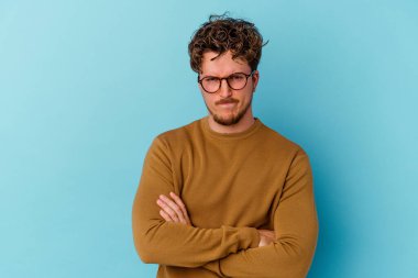 Mavi arka planda izole edilmiş gözlüklü genç beyaz adam alaycı bir ifadeyle kameraya bakıyor..
