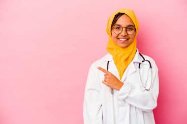 Genç doktor müslüman kadın pembe arka planda izole edilmiş gülümseyen ve işaret eden boş alanda bir şey gösteriyor..