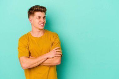 Mavi arka planda izole edilmiş genç beyaz adam kollarını kavuşturarak gülümsüyor..