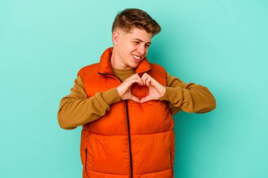 Mavi arka planda izole edilmiş genç beyaz adam gülümsüyor ve elleriyle kalp şeklini gösteriyor..